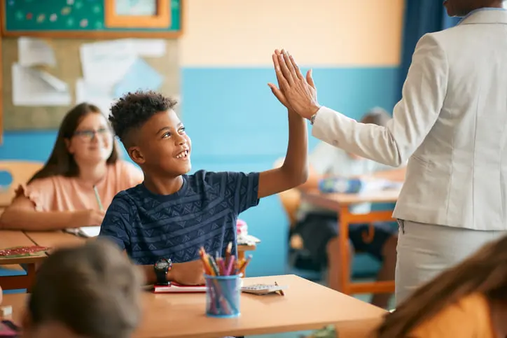 Happy Black Elementary Student And His Teacher Giving High Five During Class At School.