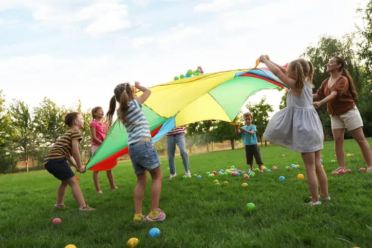 Group Of Children And Teacher Playing With Rainbow Playground Parachute On Green Grass. Summer Camp Activity